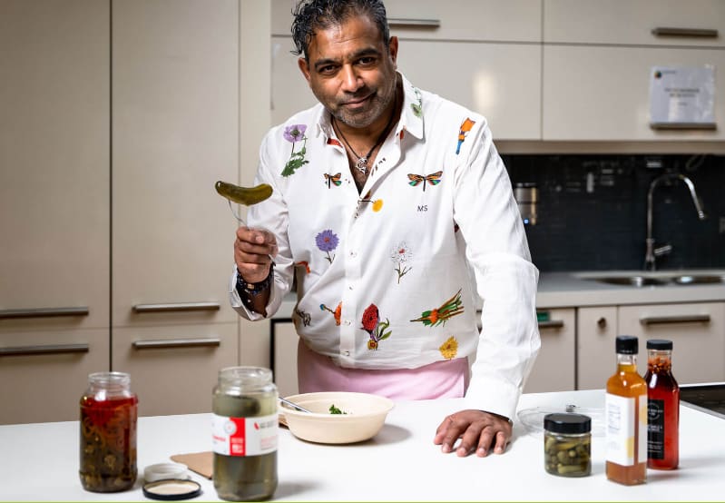 Chef in a printed jacket tasting from a spoon beside jars of preserves in a development kitchen